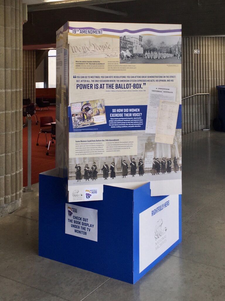 Picture of 19th Amendment Popout located near the circulation desk in the Claire T. Carney Library. The popout displays images and text related to women's right to vote.